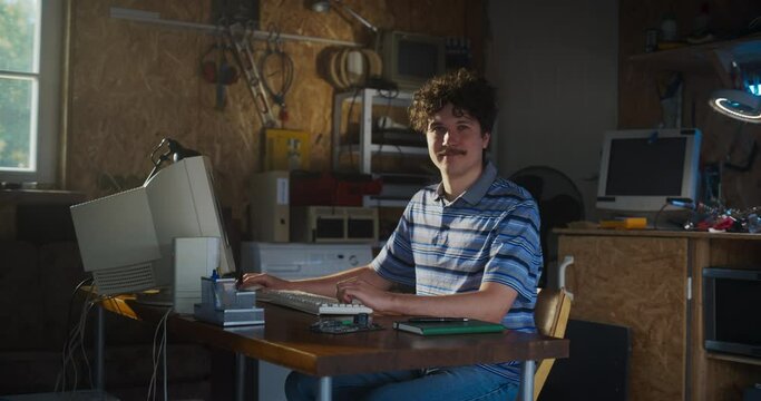 Caucasian Male Software Engineer Programming On Old Desktop Computer In Retro Garage, Looking At Camera And Smiling. Man Starting an Innovative Startup Company In Nineties. Nostalgia Concept.