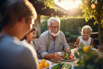 Happy Senior Grandfather Talking and Having Fun with His Grandchildren, Holding Them on Lap at a Outdoors Dinner with Food and Drinks, Adults at a Garden Party Together with Kids