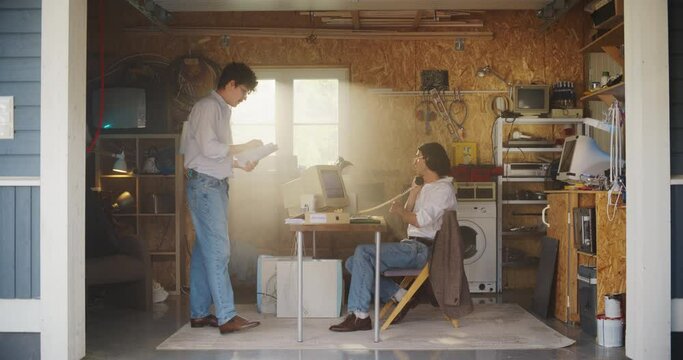 Two Caucasian Male Stock Traders Working In Retro Garage, Using Old Desktop Computer With Candlestick Chart And Landline Phone To Pitch A Public Company To Investors. Partners Starting Investment Fund