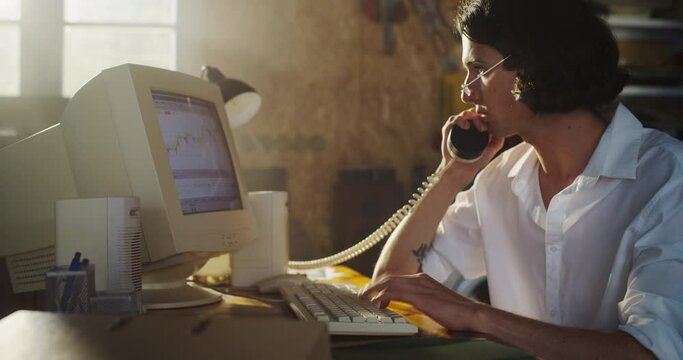 Caucasian Male Trader Talking On Landline Phone And Using Old Desktop Computer In Retro Garage. Young Man Pitching New Promising Stock To Investors. Starting Investment Management Fund in Nineties.