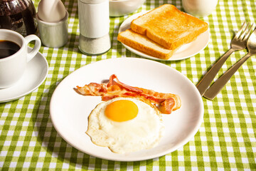 Fried egg with roasted bacon, black coffee, toasts on checkered tablecloth. Retro American breakfast. Hard shadow.