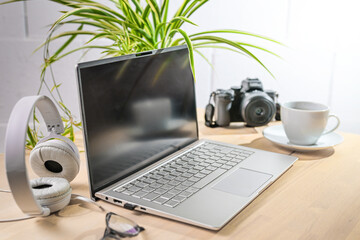 Laptop, headphones and camera on a wooden office desk with coffee cup a plant against a white wall,...