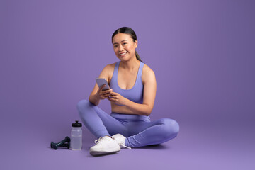 Asian woman with cellphone, resting post-exercise on purple studio background