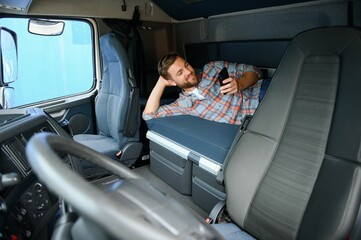 Truck driver lying on bed in his truck cabin behind driver seat and holding phone chatting with his...
