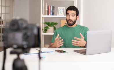 Concentrated young arabic man blogger at table with laptop shooting vlog, online lesson