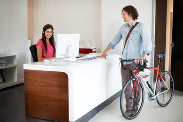 Office, desk and man with bike for business, travel and talking about commute to work with receptionist in morning. Businessman, bicycle and conversation with woman in administration or secretary