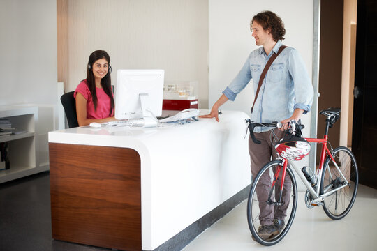 Office, desk and man with bike for business, travel and talking about commute to work with receptionist in morning. Businessman, bicycle and conversation with woman in administration or secretary
