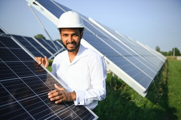 Portrait of Young indian man technician wearing white hard hat standing near solar panels against blue sky.Industrial worker solar system installation, renewable green energy generation concept.