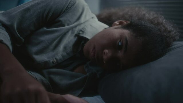 Sad African American teenage girl lying on bed at night, doomscrolling on mobile phone and then hugging her knees and closing eyes