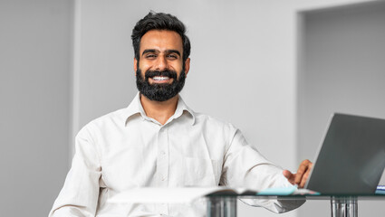 Portrait of happy indian man sitting at desk in office, working on laptop and smiling at camera, panorama, free space