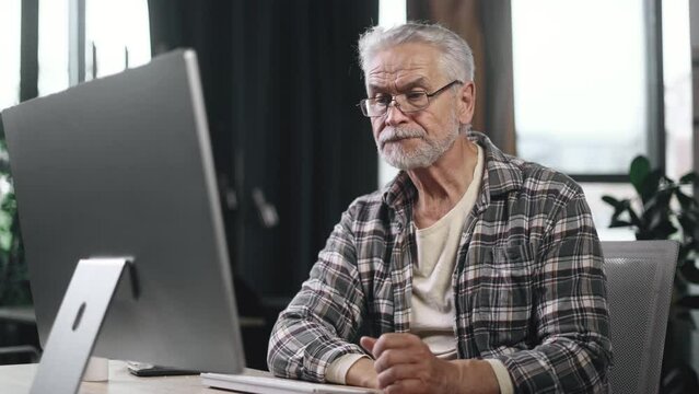 Tired boring gray haired senior man freelancer feeling annoyed at workplace Sad overworked employee mature worker falling exhausted next to computer indoors. High quality 4k footage