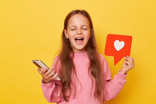 Excited amazed little girl sharing popular content on social media showing blogger heart like icon and using smartphone yelling with closed eyes rejoicing isolated over yellow background