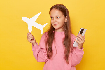 Smiling pensive little girl standing with smartphone and paper plane in hands booking tickets for...