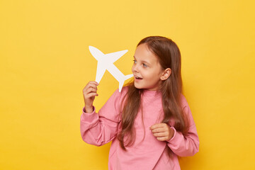 Cheerful charming little girl wearing casual clothing showing airplane and its direction dreaming to traveling having exited expression standing isolated over yellow background.