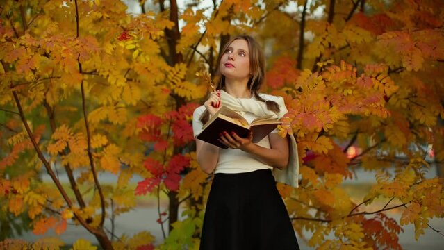 Portrait of a young girl holding a book against the background of autumn colorful leaves.