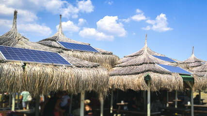 Blue tropical sky and authentic traditional Polynesian thatched roof houses with eco-friendly use of solar panels. View on the beach with a building with a solar panel on the roof. - obrazy, fototapety, plakaty