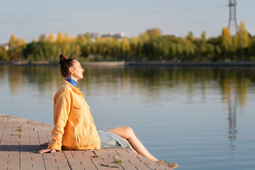 A young woman sits by the river on the embankment. Concept: freedom, enjoying life, being alone with yourself.