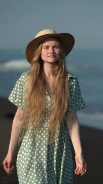 Smiling sensual blonde female wearing in straw hat, summer polka dot dress walking on beach of Pacific Ocean. 21-year-old stylish young adult model. Vertical format video, slow motion, handheld shot