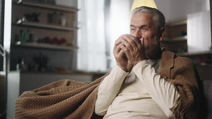 Portrait of handsome gray haired senior man under the blanket take sip drinking hot tea or coffee sitting on couch at home Calm relaxed mature bearded male enjoying weekend leisure time alone indoors