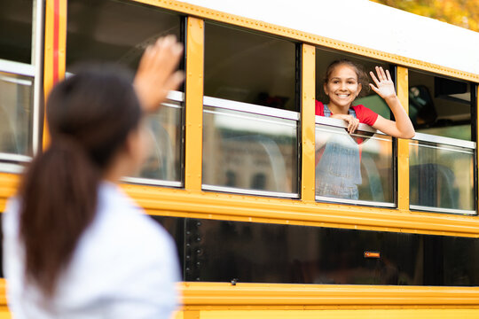 Young mom waving goodbye to happy daughter sitting in yellow school bus