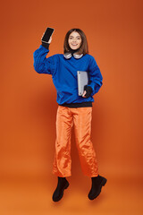 Fototapeta premium cheerful woman in trendy autumnal outfit jumping with gadgets on orange, cyber monday concept