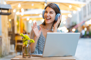 Happy relaxed overjoyed pretty young woman in wireless headphones choosing listening favorite energetic disco music in laptop computer. Girl sits in urban city cafe restaurant terrace dancing outdoors