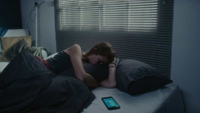 Young woman lying in bed at night, reading embarrassing message on smartphone, hugging pillow and crying