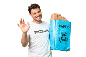 Brazilian man holding a recycling bag full of paper to recycle over isolated chroma key background saluting with hand with happy expression