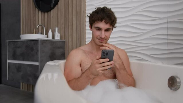 Caucasian pensive calm tranquil man naked relaxed guy male sitting relax in bathtub in soapy foam warm hot water bath thoughtful looking mobile phone using smartphone think reading social media app