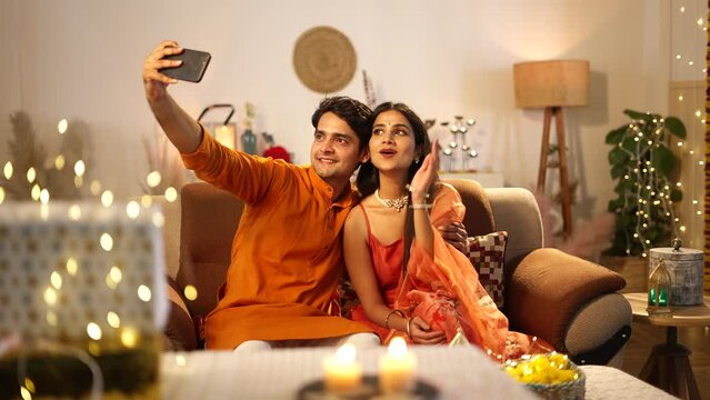 Indian beautiful couple click a selfie picture while sitting on the sofa.wife is doing flying kiss,well decorated home,hindu festival concept.
