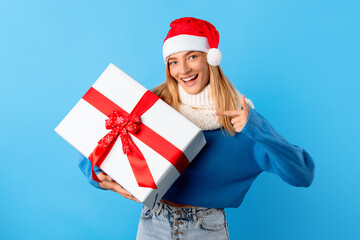 Happy european lady in Santa hat holding wrapped present box and pointing finger on it, standing over blue background