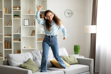 Domestic entertainments. Joyful woman in wireless headphones enjoying music and dancing on sofa to favorite song, living room, copy space