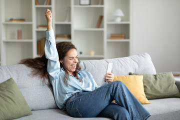 Overjoyed young woman holding smartphone and shaking fist in joy, reading great news and message,...