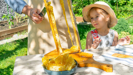 A girl eats honey in a honeycomb. A girl in a hat watches her grandfather cut fresh honey into a...