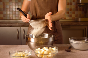 woman chef in brown apron sifts flour through sieve for making dough on cozy kitchen background
