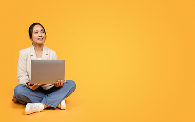 Positive korean millennial lady in suit typing on laptop, sit on floor, look at copy space