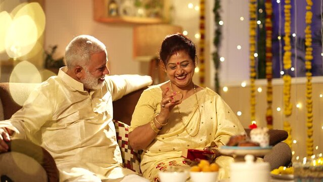 Handsome Indian aged man gifting gold bangles to his beautiful wife on the Diwali festival.they both are sitting on the sofa.looking good in traditional dress,decorated home.