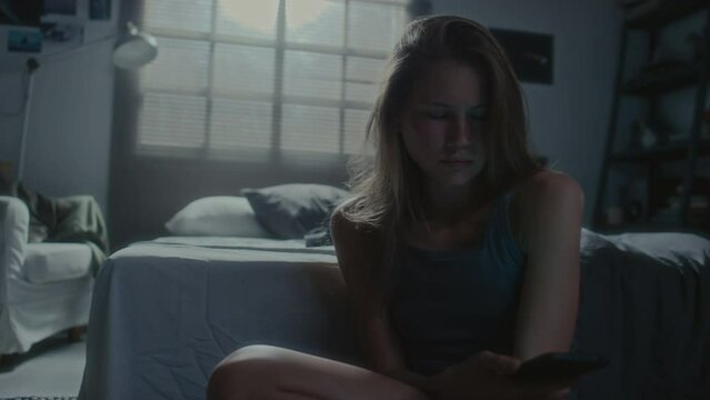 Handheld shot of sad teen girl sitting on floor in dark bedroom, reading abusive message, putting phone away and hugging knees while being cyberbullied