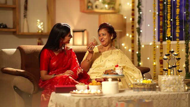 Beautiful Indian woman  giving a gold bangle Diwali gift to her mother- in- law.they both are wearing traditional dress.they both are very happy and sitting on the sofa.festive mood at home.