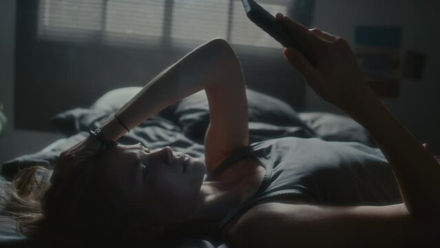 Young woman lying on bed, reading humiliating messages on phone, getting upset and turning on side during sleepless night