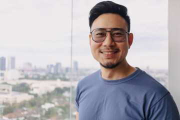 Asian man wear glasses and blue t-shirt with beard, smiling and standing over city view at condominium, looking at camera at apartment room alone, having happy good life.