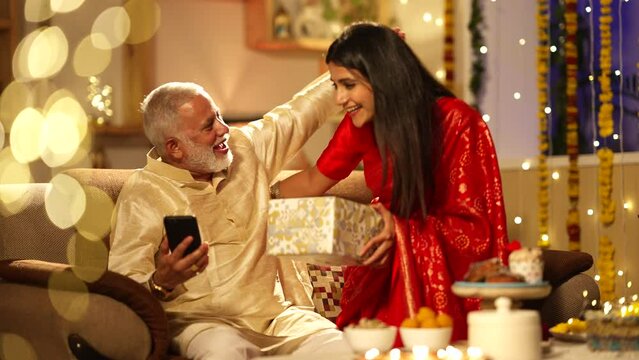 Indian daughter giving a diwali gift to her father while cheking the mobile phone.they both are sitting on the sofa.Diwali decorations,wearing traditional dress.festival concept.