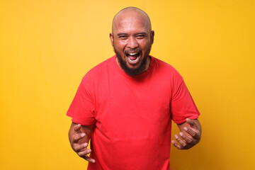Bald bearded fat man in red t-shirt shouting screaming with eyes closed over yellow background