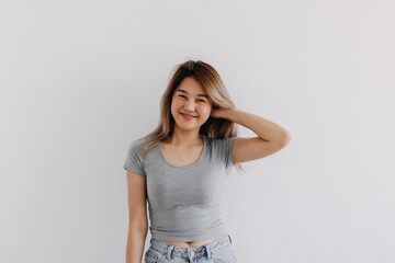 Happy Asian Thai woman wear grey, smiling and raising her hand up for touching and grabbing hair, looking at empty camera, chilling standing isolated on white background wall.