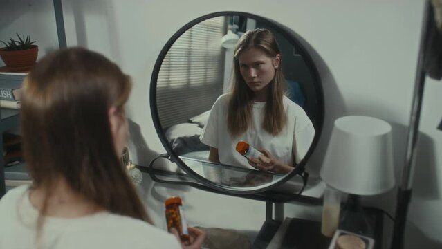 Young depressed girl sitting at home, holding pills and looking at her reflection in mirror while thinking about suicide