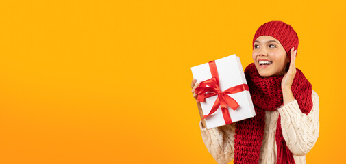 Joyful lady holding gift holding hand near ear, yellow background