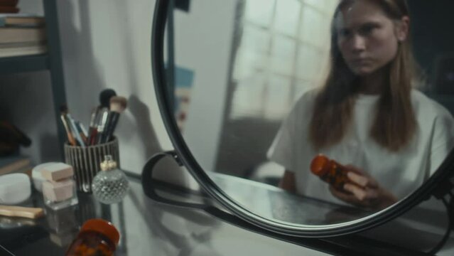 Tilt up shot of depressed girl holding pills, looking at her reflection in mirror and thinking about suicide while sitting at home