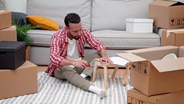 A young man assembles a DIY shelf amid moving carton boxes, showcasing his resourcefulness in his new home after relocating