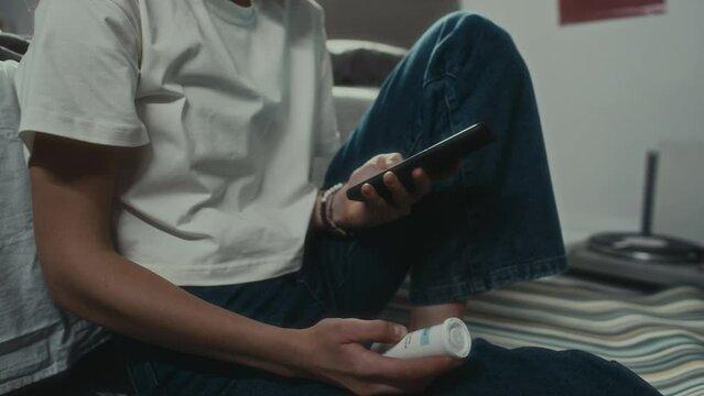 Tilt up shot of frustrated girl sitting on floor in bedroom, reading messages on phone and holding pills while having suicidal thoughtsTilt up shot of frustrated girl sitting on floor in bedroom, read