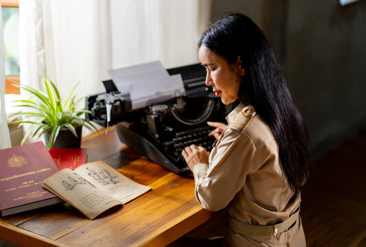 Asian woman wear uniform look like teacher of goverment worker work with typewriter and type the detail from old book in front of window and warm light.